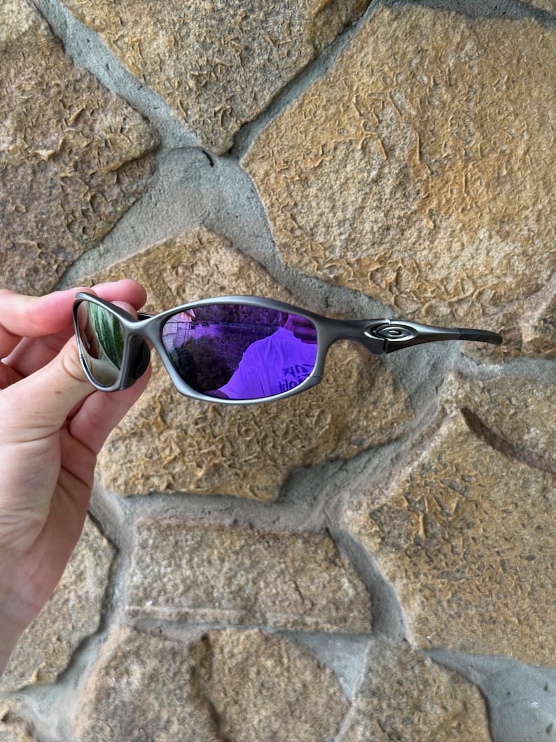May include: A pair of silver-framed sunglasses with purple mirrored lenses. The sunglasses have a sleek, modern design with a curved frame and dark temples. The lenses reflect the surrounding environment, creating a vibrant purple hue.