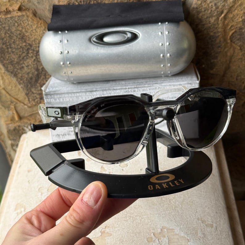 May include: Clear-framed Oakley sunglasses with dark lenses rest on a black display stand. The stand has the Oakley logo. A silver case and black cloth are in the background.