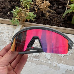 May include: Gray sports sunglasses with a vibrant pink and orange mirrored lens. The sunglasses have a sleek, modern design with a gray frame and arms. The lenses reflect a gradient of colors, adding a stylish touch.