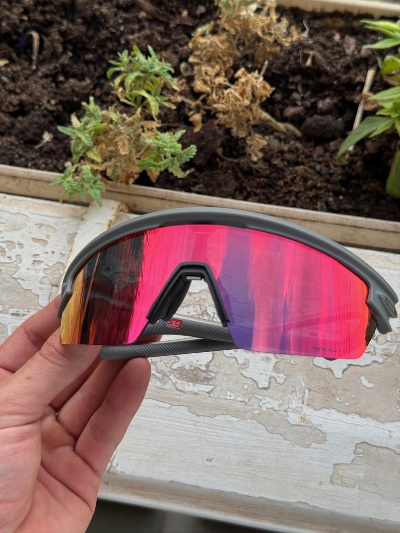 May include: Gray sports sunglasses with a vibrant pink and orange mirrored lens. The sunglasses have a sleek, modern design with a gray frame and arms. The lenses reflect a gradient of colors, adding a stylish touch.