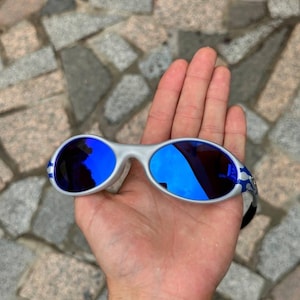 May include: Silver-framed sunglasses with blue mirrored lenses. The sunglasses have a curved design and blue accents on the temples. The lenses reflect the surrounding environment. The sunglasses are held in an open hand.