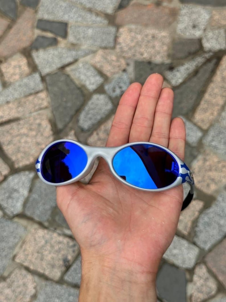 May include: Silver-framed sunglasses with blue mirrored lenses. The sunglasses have a curved design and blue accents on the temples. The lenses reflect the surrounding environment. The sunglasses are held in an open hand.