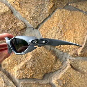 May include: Gray sunglasses with a unique, angular design. The lenses are dark, and the frame features a metallic finish. The Oakley logo is visible on the temple. These sunglasses are designed for outdoor activities.