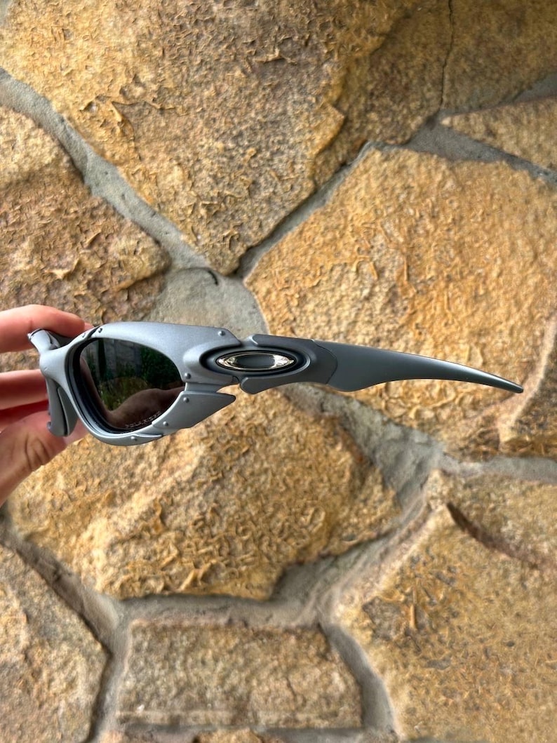 May include: Gray sunglasses with a unique, angular design. The lenses are dark, and the frame features a metallic finish. The Oakley logo is visible on the temple. These sunglasses are designed for outdoor activities.