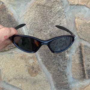 May include: Black sunglasses with a curved frame and dark lenses. The sunglasses have a sleek, sporty design, with a small white logo on the temples. The lenses reflect the surrounding environment.