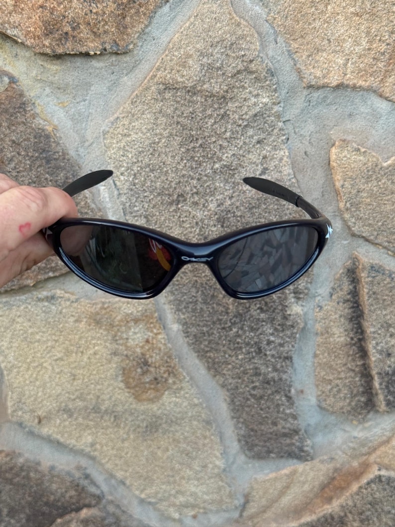 May include: Black sunglasses with a curved frame and dark lenses. The sunglasses have a sleek, sporty design, with a small white logo on the temples. The lenses reflect the surrounding environment.