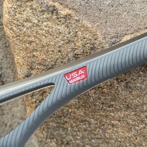 May include: Close-up of a gray grilling tool with a ridged handle. A red rectangular label with the text "USA ASSEMBLED" is visible. The tool is set against a textured, light brown stone background.