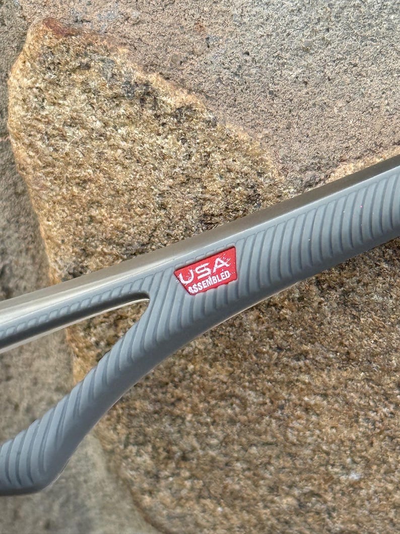 May include: Close-up of a gray grilling tool with a ridged handle. A red rectangular label with the text "USA ASSEMBLED" is visible. The tool is set against a textured, light brown stone background.