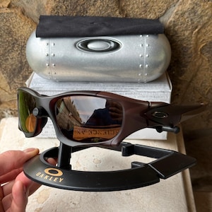 Może przedstawiać: Brązowe i czarne okulary przeciwsłoneczne Oakley z odblaskowymi soczewkami, prezentowane na czarnym stojaku ze złotym logo Oakley. Srebrne etui z logo Oakley i czarna ściereczka znajdują się w tle.