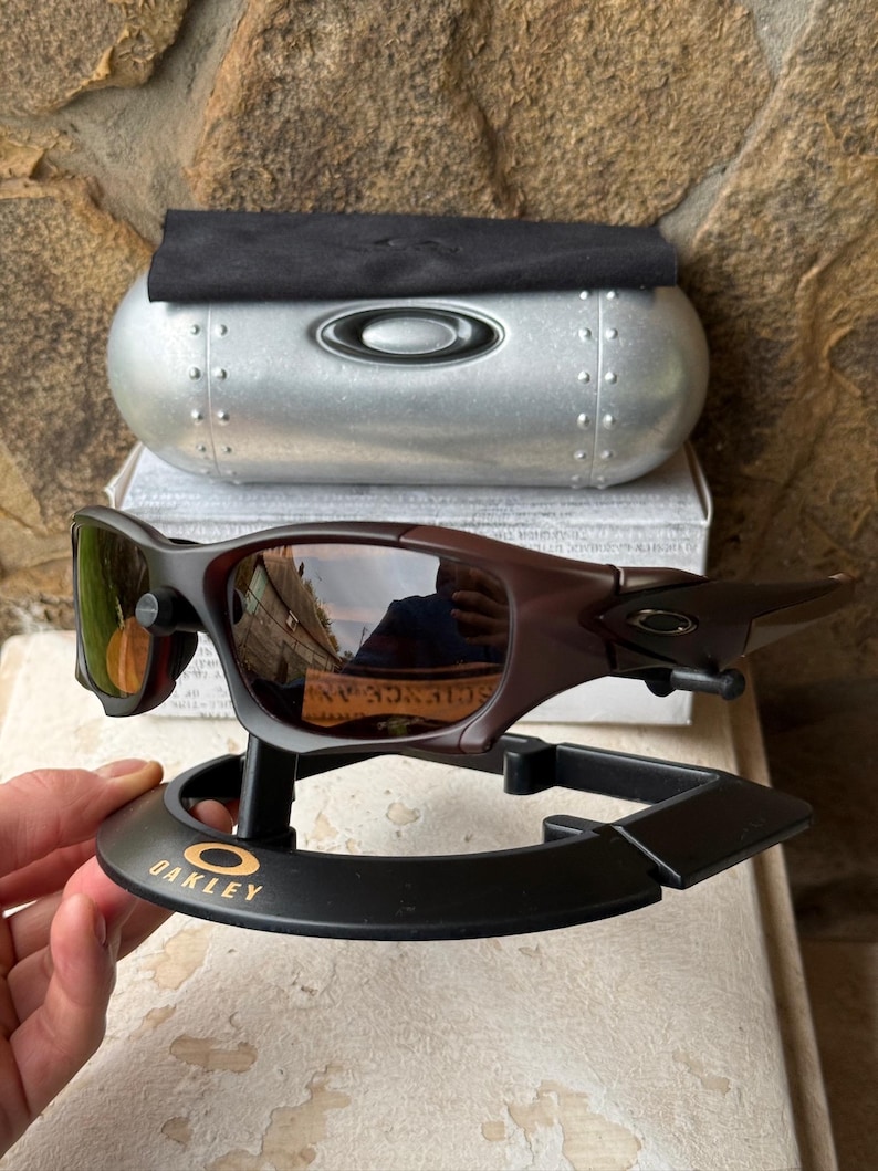 Może przedstawiać: Brązowe i czarne okulary przeciwsłoneczne Oakley z odblaskowymi soczewkami, prezentowane na czarnym stojaku ze złotym logo Oakley. Srebrne etui z logo Oakley i czarna ściereczka znajdują się w tle.