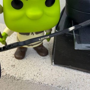 May include: A green Shrek Funko Pop figure with brown boots and a beige shirt. The word "EJECTOR" is printed on a black object in the foreground. A black box is in the background.