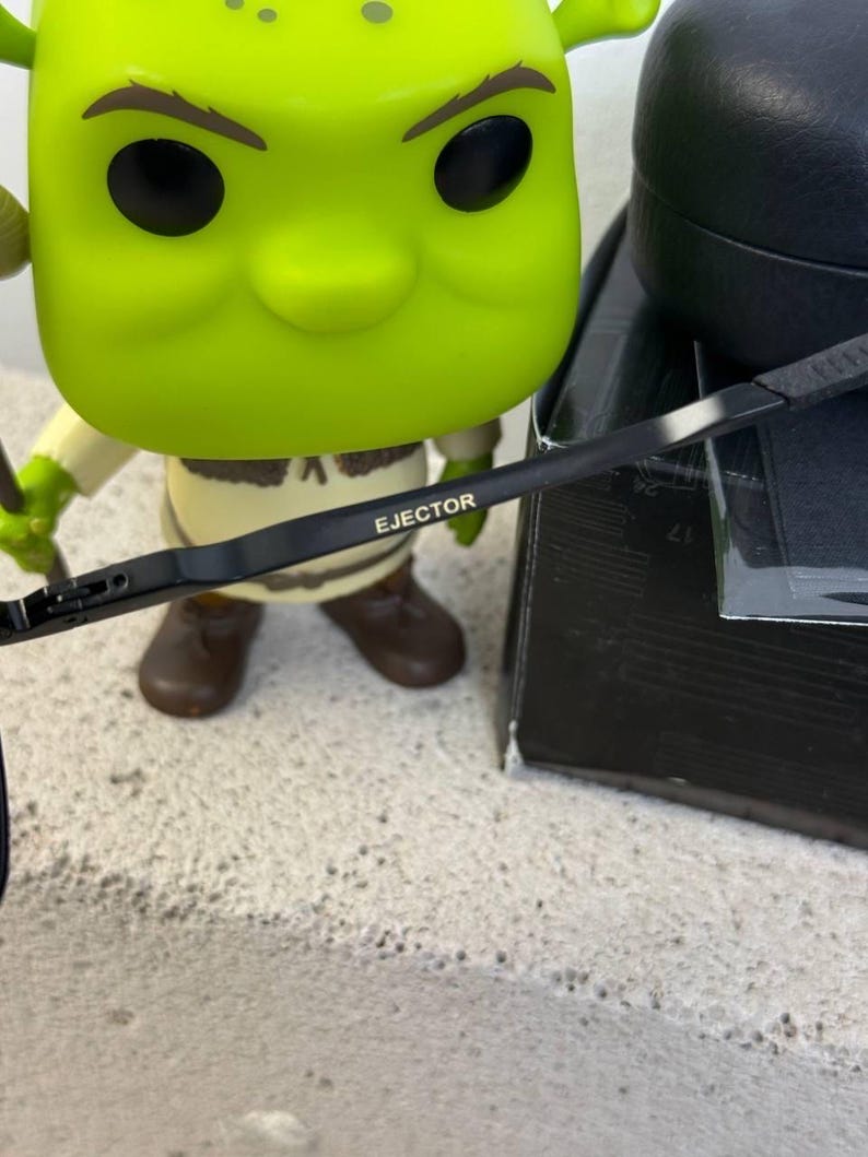May include: A green Shrek Funko Pop figure with brown boots and a beige shirt. The word "EJECTOR" is printed on a black object in the foreground. A black box is in the background.