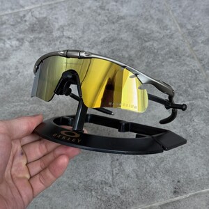 May include: Sport sunglasses with a metallic gray frame and mirrored yellow lenses. The sunglasses are displayed on a black stand with the "OAKLEY" logo. The lenses reflect the surrounding environment.