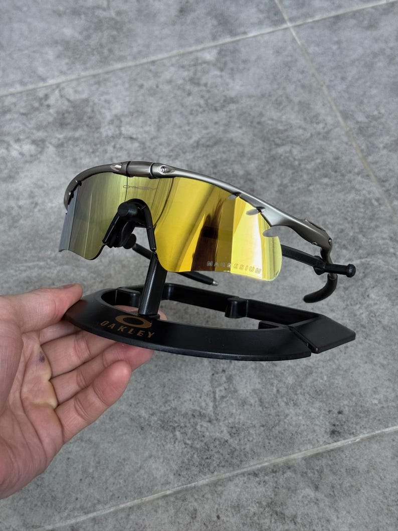 May include: Sport sunglasses with a metallic gray frame and mirrored yellow lenses. The sunglasses are displayed on a black stand with the "OAKLEY" logo. The lenses reflect the surrounding environment.