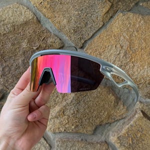 May include: Gray sports sunglasses with mirrored pink and orange lenses. The sunglasses have a sleek, modern design with a black nose bridge and gray frames. The lenses reflect a vibrant array of colors.