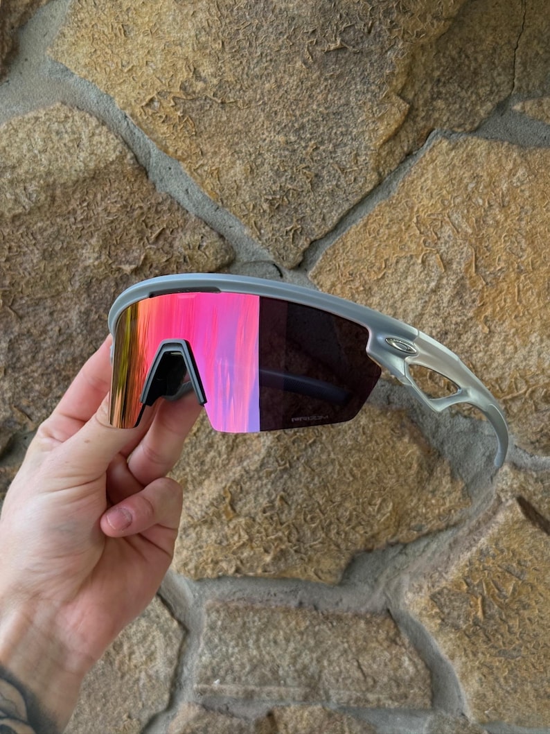 May include: Gray sports sunglasses with mirrored pink and orange lenses. The sunglasses have a sleek, modern design with a black nose bridge and gray frames. The lenses reflect a vibrant array of colors.