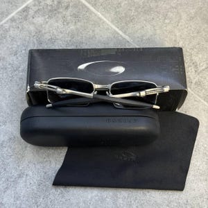 May include: Silver-framed sunglasses with dark lenses sit on a black case and a black microfiber cloth. The case and cloth have the brand name "Oakley" printed on them. A black box with the Oakley logo is behind the sunglasses.