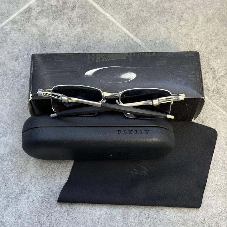 May include: Silver-framed sunglasses with dark lenses sit on a black case and a black microfiber cloth. The case and cloth have the brand name "Oakley" printed on them. A black box with the Oakley logo is behind the sunglasses.