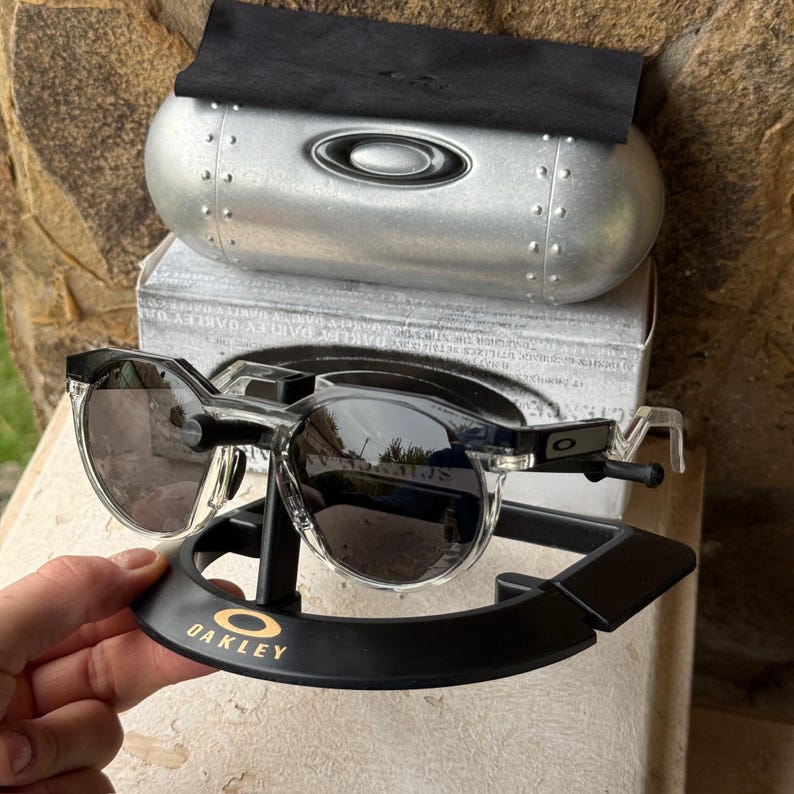 May include: Clear-framed sunglasses with dark lenses rest on a black display stand with the gold "Oakley" logo. The sunglasses are paired with a silver case and a black microfiber cloth. The lenses reflect the surrounding environment.