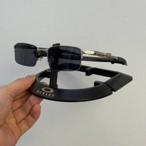 May include: Dark sunglasses with a silver frame are displayed on a black stand. The stand features the gold "OAKLEY" logo. The sunglasses have rectangular lenses and are designed for eye protection.