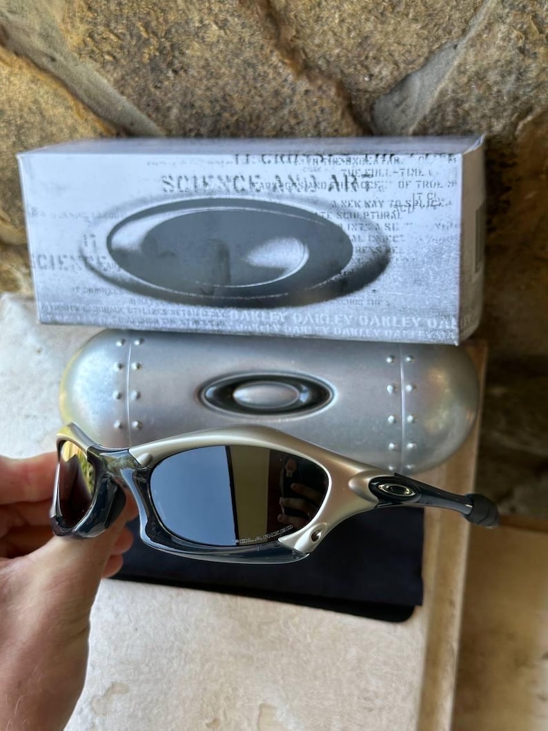 May include: Silver and black sunglasses with dark lenses, a silver case, and a box with the text "SCIENCE AND ART". The sunglasses have a sleek, modern design.