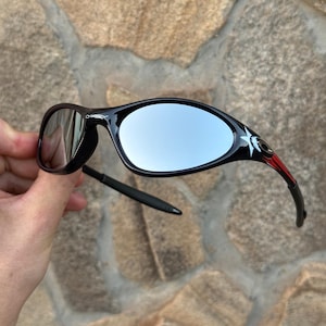 May include: Black sunglasses with mirrored lenses. The frames are black with red and white accents near the temples. The lenses have a reflective, silver appearance. The sunglasses are held against a stone background.