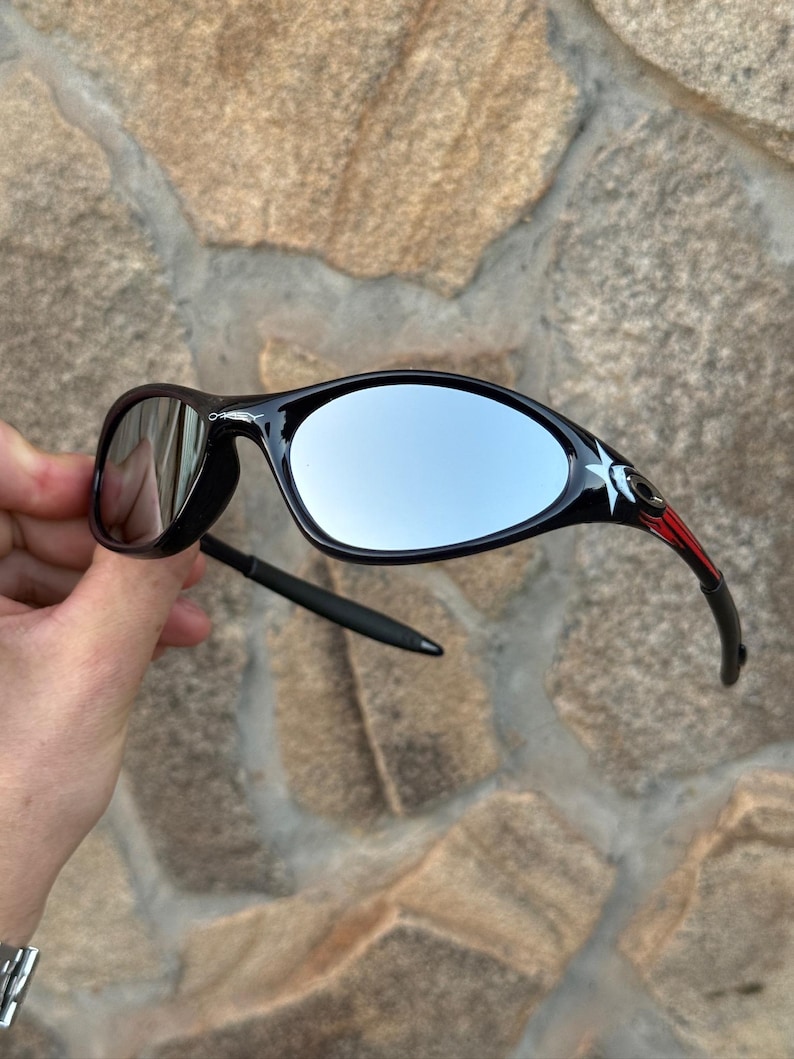 May include: Black sunglasses with mirrored lenses. The frames are black with red and white accents near the temples. The lenses have a reflective, silver appearance. The sunglasses are held against a stone background.