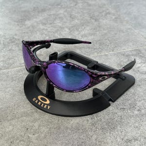 Puede incluir: Gafas de sol Oakley moradas y negras con un patr&oacute;n veteado. Las lentes tienen un degradado de azul a morado. Las gafas de sol se exhiben en un soporte negro con el logotipo de Oakley.