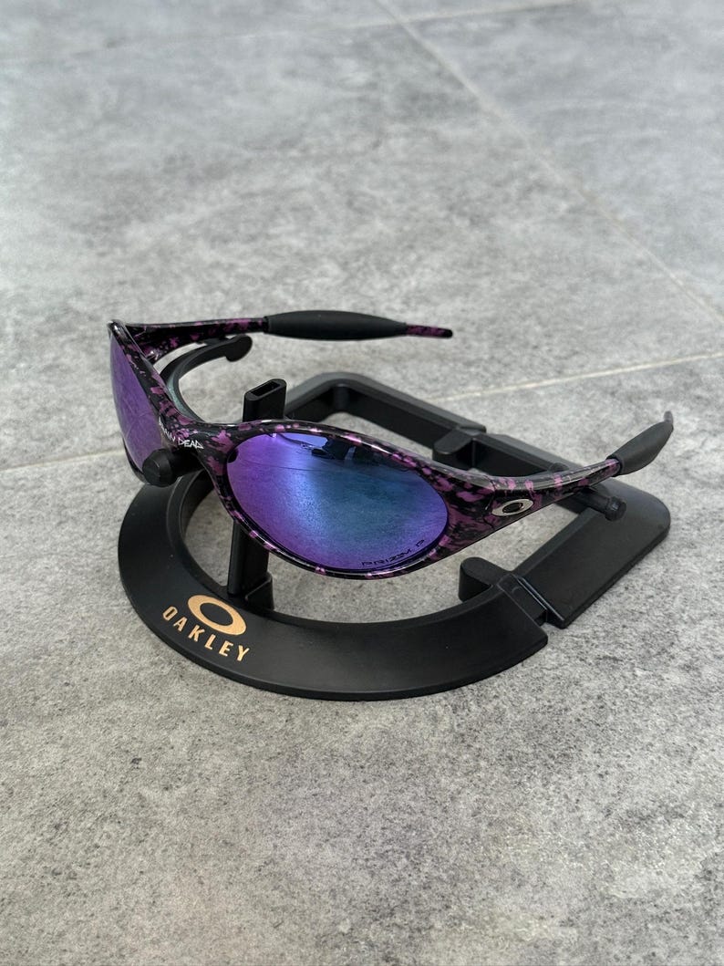 Puede incluir: Gafas de sol Oakley moradas y negras con un patr&oacute;n veteado. Las lentes tienen un degradado de azul a morado. Las gafas de sol se exhiben en un soporte negro con el logotipo de Oakley.