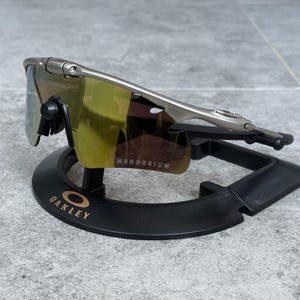 May include: Silver-framed Oakley sunglasses with yellow-tinted lenses. The lenses have a mirrored finish and the word "MAGNESIUM" is printed on the lens. The sunglasses are displayed on a black stand with the Oakley logo.