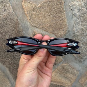 May include: Black sunglasses with a red accent on the frame. The sunglasses have a sleek, modern design with dark lenses and a curved shape. The arms are black and the lenses are dark, providing a stylish look.