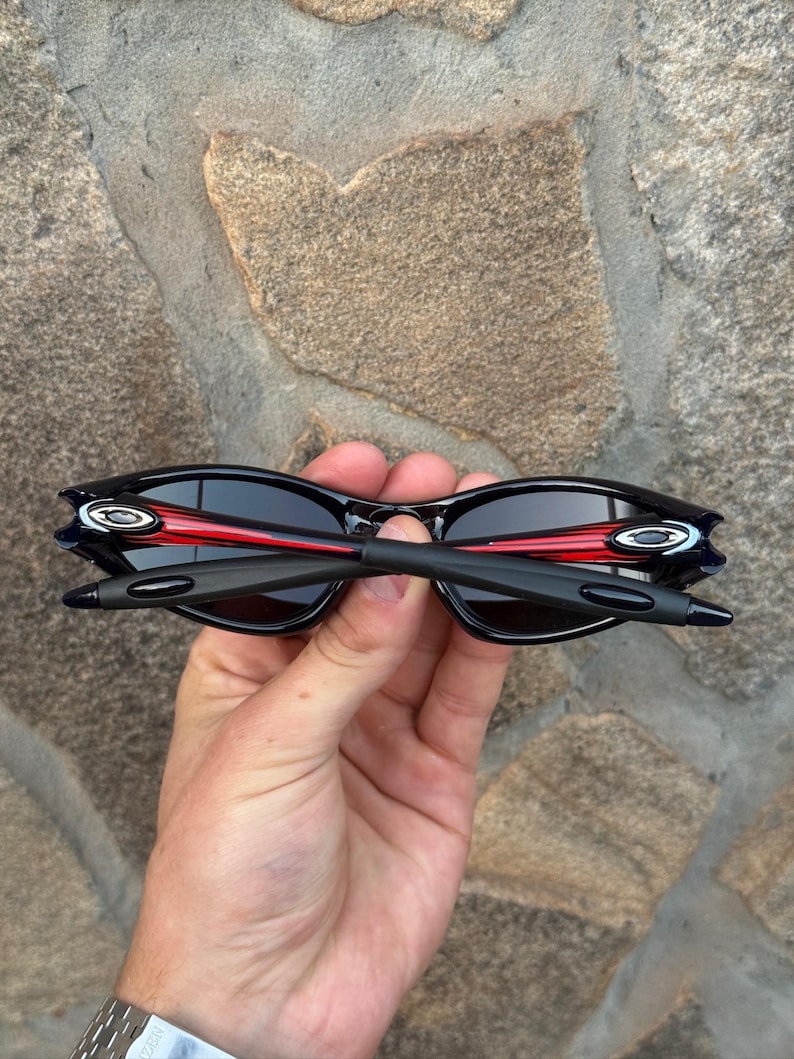 May include: Black sunglasses with a red accent on the frame. The sunglasses have a sleek, modern design with dark lenses and a curved shape. The arms are black and the lenses are dark, providing a stylish look.