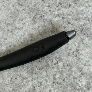 May include: Close-up of a black stylus with a silver tip. The stylus has a matte finish and a rounded shape. The letters "CE" are imprinted on the side of the stylus.