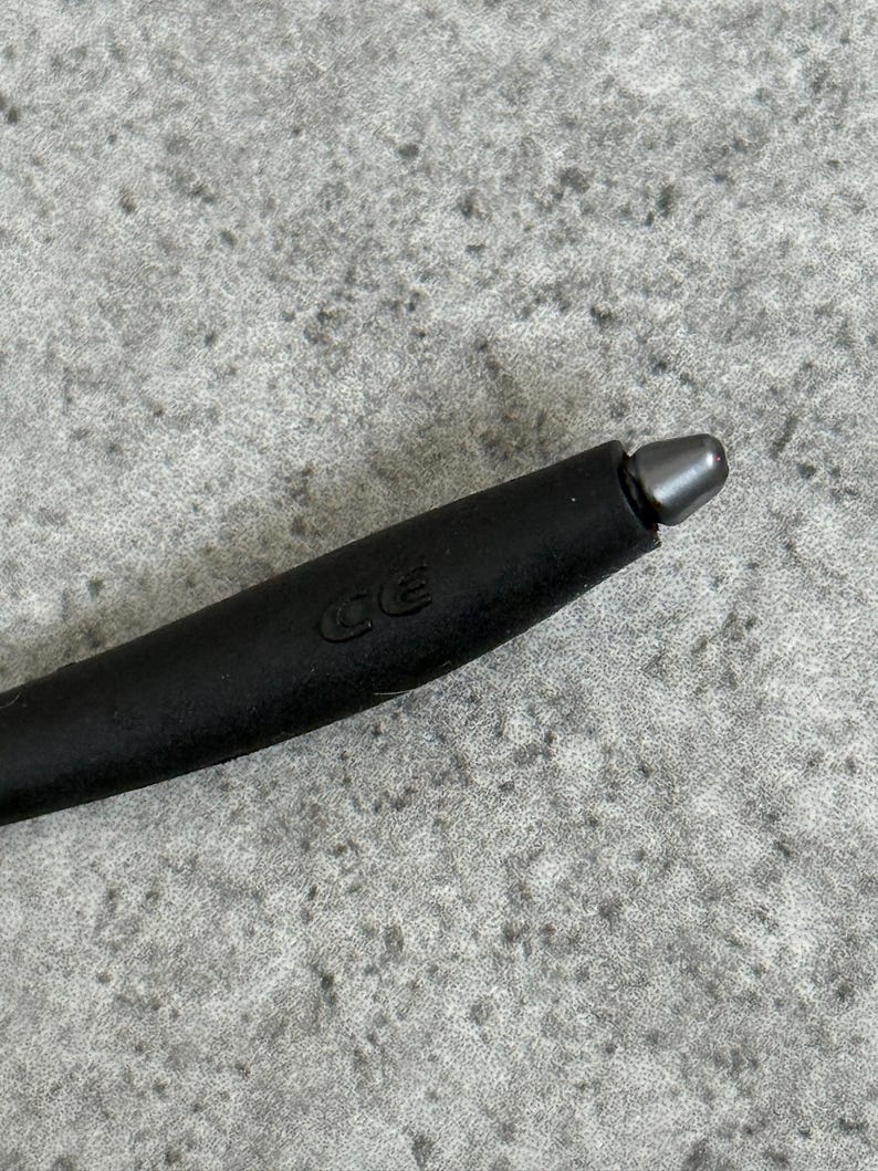 May include: Close-up of a black stylus with a silver tip. The stylus has a matte finish and a rounded shape. The letters "CE" are imprinted on the side of the stylus.