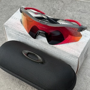 May include: Performance sunglasses featuring a single-piece wrap-around red and orange mirrored lens, a black frame, and red temple tips. The eyewear is positioned on a white box with text, accompanied by a black textured hard-shell carrying case.