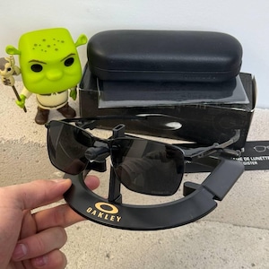 May include: Black Oakley sunglasses with a square frame, displayed on a black stand with the Oakley logo in gold. The sunglasses are next to a black case and a box. A green Shrek figurine is also in the image.
