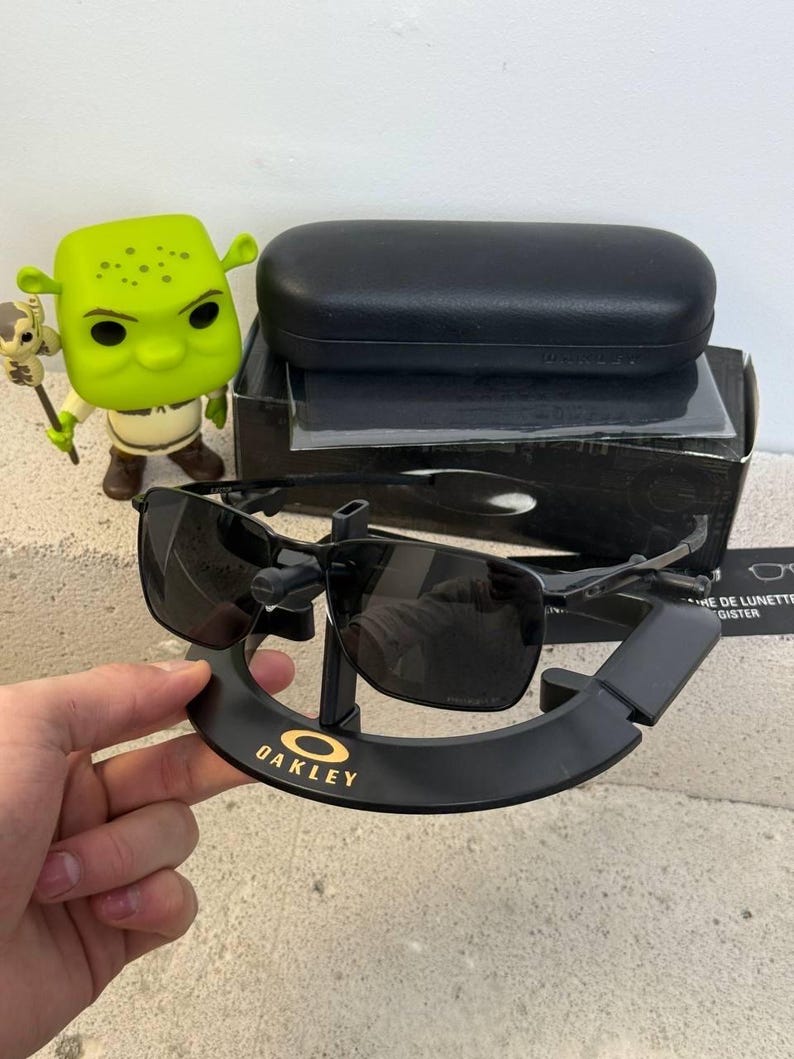 May include: Black Oakley sunglasses with a square frame, displayed on a black stand with the Oakley logo in gold. The sunglasses are next to a black case and a box. A green Shrek figurine is also in the image.