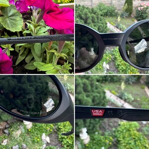 K&ouml;nnte beinhalten: Schwarze Oakley-Sonnenbrille mit dunklen Gl&auml;sern. Die Sonnenbrille hat ein abgerundetes Rahmendesign. Der Text "USA Assembled" ist auf dem B&uuml;gel zu sehen. Die Sonnenbrille befindet sich vor einem Hintergrund aus rosa Blumen und Gr&uuml;n.