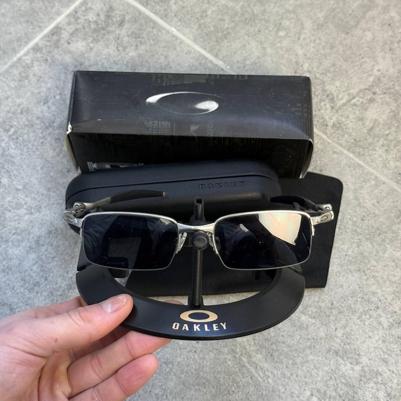 May include: Silver-framed Oakley sunglasses with dark lenses, presented on a black stand bearing the Oakley logo. The rectangular sunglasses are accompanied by a black case and box.