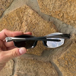 May include: Black sunglasses with a silver frame and dark lenses. The sunglasses are held against a textured stone background. The arms of the sunglasses are thin and sleek, with a silver accent near the hinges.