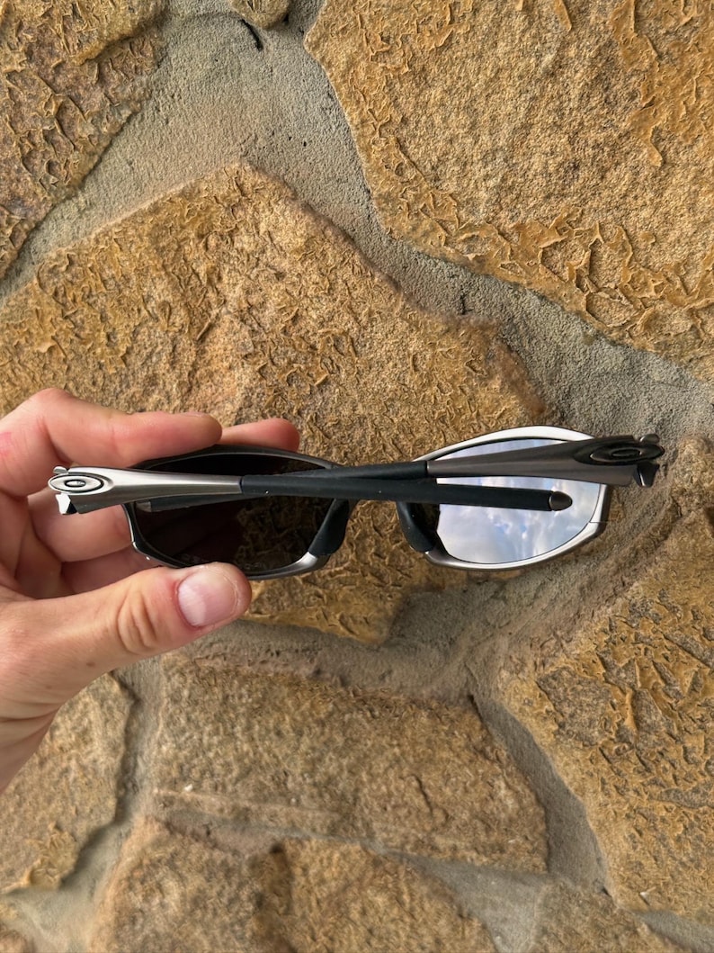 May include: Black sunglasses with a silver frame and dark lenses. The sunglasses are held against a textured stone background. The arms of the sunglasses are thin and sleek, with a silver accent near the hinges.