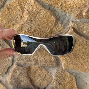 May include: Silver-framed sunglasses with dark lenses. The sunglasses have a curved design and are held against a stone background. A silver case with the Oakley logo is visible below the sunglasses.