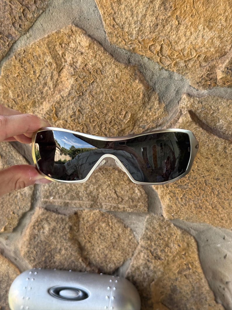 May include: Silver-framed sunglasses with dark lenses. The sunglasses have a curved design and are held against a stone background. A silver case with the Oakley logo is visible below the sunglasses.