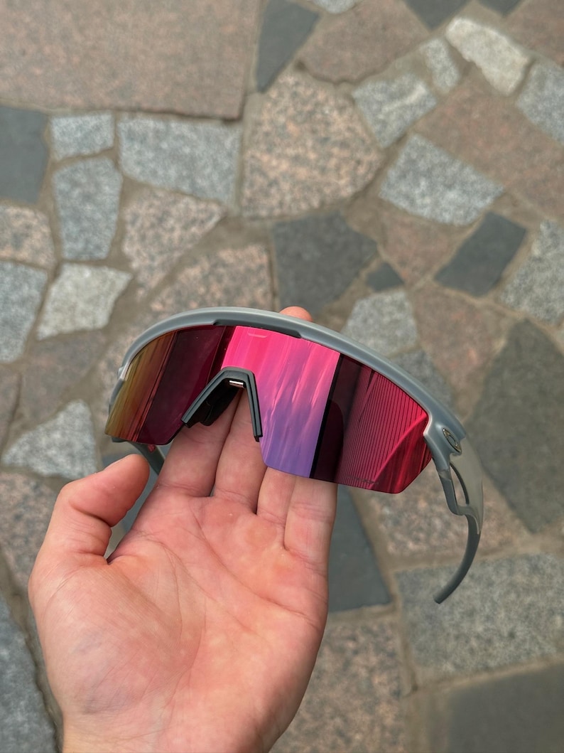 May include: Gray sports sunglasses with red mirrored lenses. The sunglasses have a sleek, modern design with a gray frame and curved temples. The lenses reflect a gradient of red and purple hues.