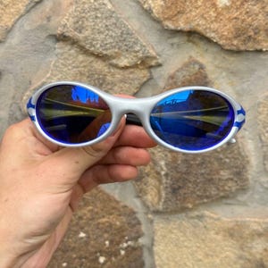 May include: Silver-framed sunglasses with blue lenses and blue and white accents on the temples. The sunglasses have a sporty, wraparound design. The lenses reflect a blue hue.
