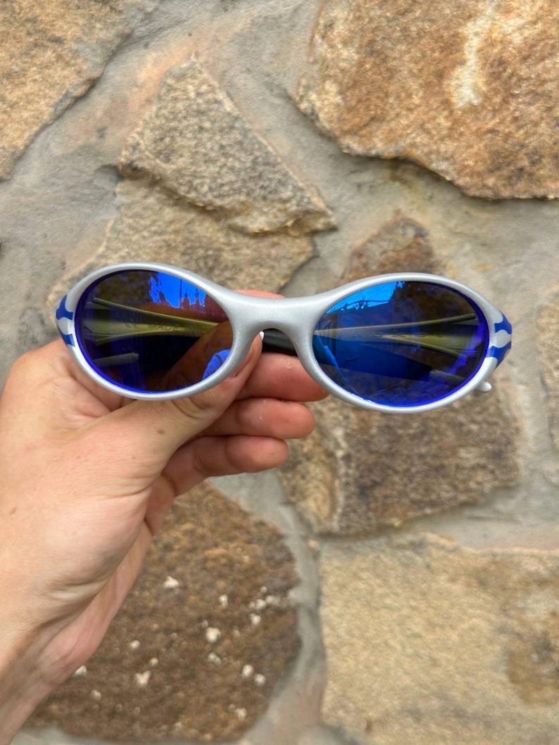 May include: Silver-framed sunglasses with blue lenses and blue and white accents on the temples. The sunglasses have a sporty, wraparound design. The lenses reflect a blue hue.