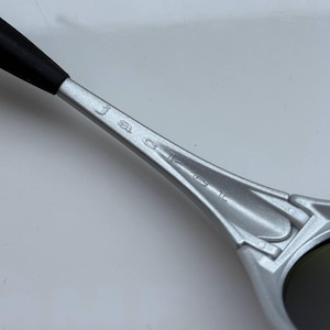 May include: Close-up of a silver-colored sunglass arm with the word "Jacket" engraved on it. The arm has a black rubber tip and a sleek, modern design. The lens is dark and reflective.