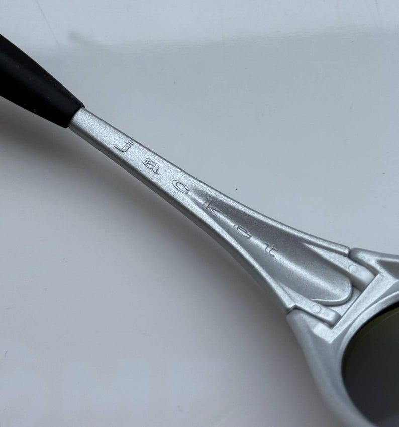 May include: Close-up of a silver-colored sunglass arm with the word "Jacket" engraved on it. The arm has a black rubber tip and a sleek, modern design. The lens is dark and reflective.