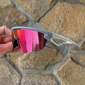 May include: Gray sports sunglasses with a vibrant pink mirrored lens. The sunglasses have a sleek, modern design with a gray frame and arms. The lenses reflect a bright pink color, offering a stylish and functional accessory for outdoor activities.