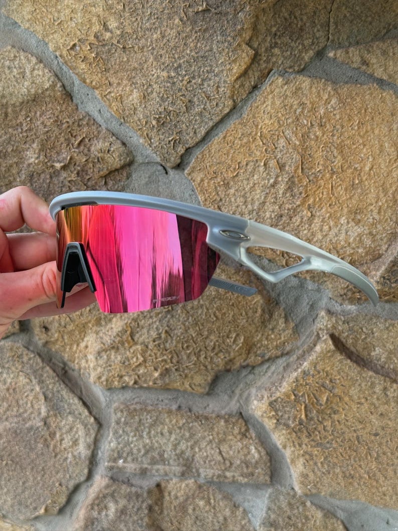 May include: Gray sports sunglasses with a vibrant pink mirrored lens. The sunglasses have a sleek, modern design with a gray frame and arms. The lenses reflect a bright pink color, offering a stylish and functional accessory for outdoor activities.
